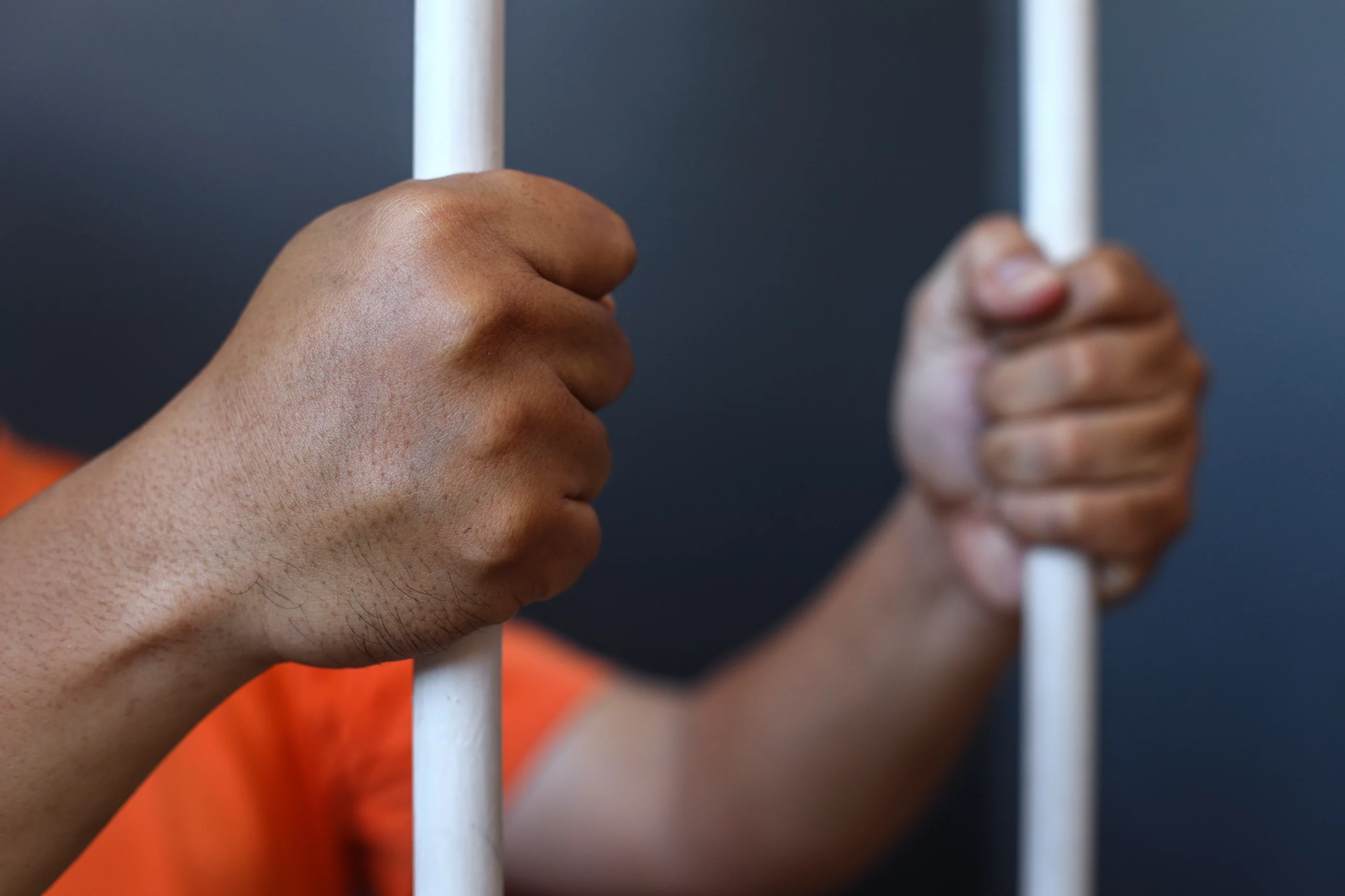 Hands gripping white jail bars, person in orange shirt — symbolizing confinement and criminal charges.